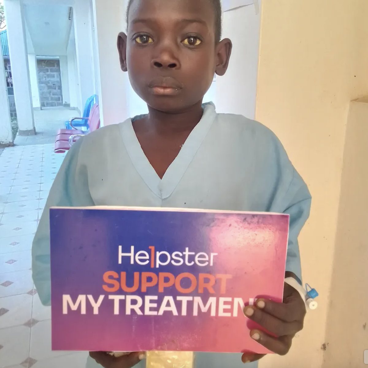 Joseph, 10, in a hospital gown, holding a Helpster sign that reads Support my treatment, in the corridor of Awasi Catholic Mission Health Centre